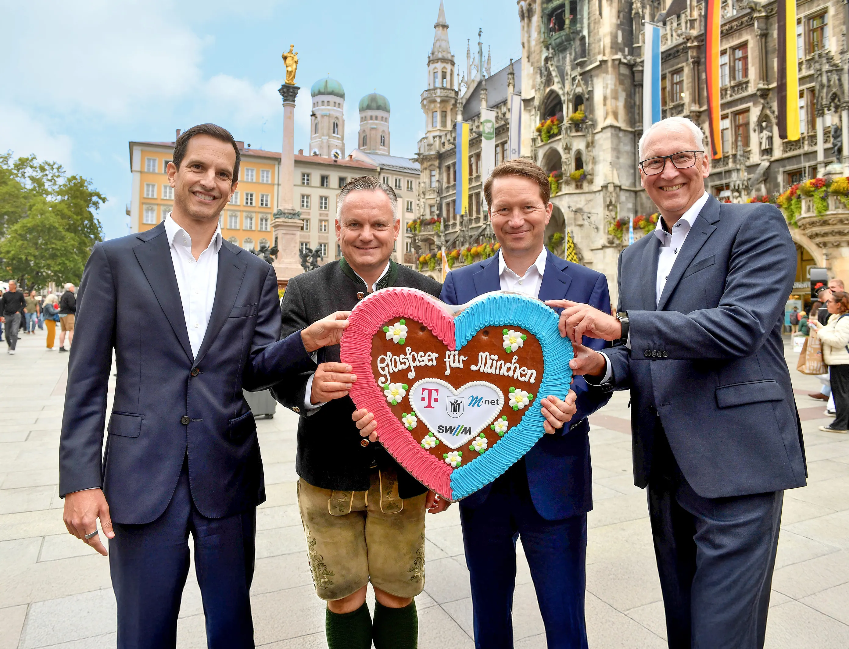 Die Kooperationspartner in Person von Rodrigo Diehl, Dr. Christian Scharpf, Dr. Florian Bieberbach, Maximilian Oertle halten gemeinsam auf dem Marienplatz ein großes Lebkuchenherz mit der Aufschrift „Glasfaser für München“ sowie den Logos von Telekom, M-net und SWM.