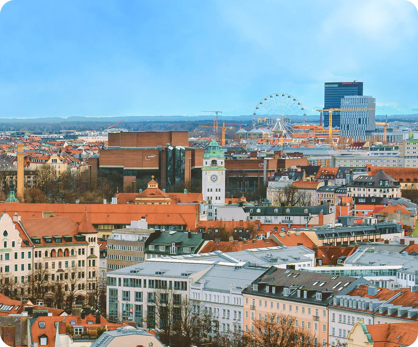 Stadtpanorama von München mit dicht bebauten Häusern und roten Ziegeldächern; im Vordergrund historische Gebäude und ein heller Uhrturm mit grünlicher Kuppel, im Hintergrund ein Riesenrad, moderne Hochhäuser und Baukräne unter klarem Himmel.